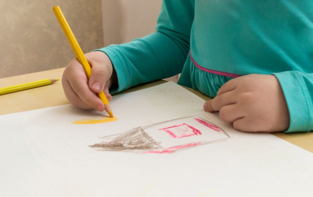 A child dressed in turquoise draws a house with colored pencils on paper indoors.